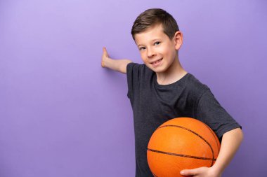 Mor arka planda izole edilmiş basketbol oynayan küçük çocuk, davet edilmek için ellerini yana uzatıyor.