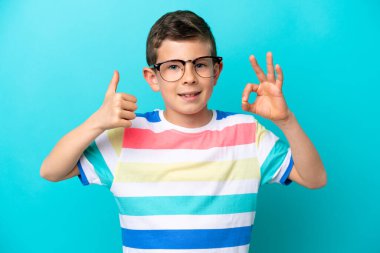 Küçük çocuk mavi arka planda gözlüklü ve iyi bir işaret yapıyor.