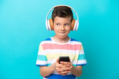 Küçük çocuk mavi arka planda izole edilmiş. Cep telefonuyla müzik dinliyor ve düşünüyor.