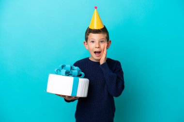 Elinde doğum günü pastası tutan küçük çocuk mavi arka planda şaşırmış ve şok olmuş bir yüz ifadesiyle izole edilmiş.