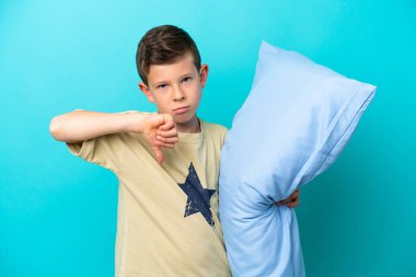 Mavi arka planda izole edilmiş pijamalı küçük bir çocuk negatif ifadeyle baş parmağını aşağı gösteriyor.