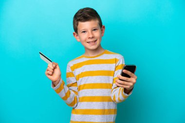 Küçük çocuk mavi arka planda izole edilmiş, kredi kartıyla cep telefonuyla alışveriş yapıyor.