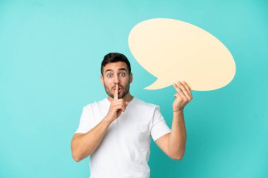 Mavi arka planda izole edilmiş yakışıklı beyaz bir adam elinde boş bir konuşma balonu ve sessizlik hareketi yapıyor.