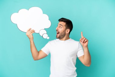 Genç, beyaz, mavi arka planda izole edilmiş genç bir adam elinde şaşırtıcı bir ifadeyle düşünen bir konuşma balonu tutuyor.