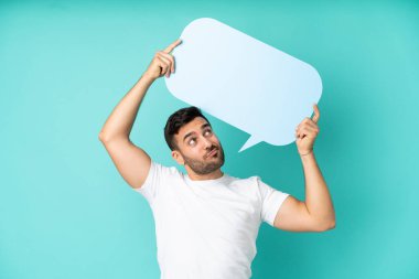 Mavi arka planda izole edilmiş yakışıklı beyaz bir adam elinde boş bir konuşma balonu ve üzgün bir ifadeyle