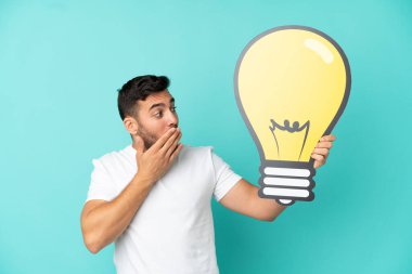 Mavi arka planda izole edilmiş yakışıklı beyaz bir adam elinde bir ampul simgesi tutuyor.