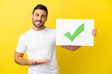 Sarı arka planda izole edilmiş yakışıklı beyaz bir adam elinde yeşil işaret simgesi olan bir pankart tutuyor ve onu işaret ediyor.
