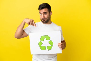 Sarı arka planda izole edilmiş yakışıklı beyaz adam elinde geri dönüşüm simgesi olan bir pankart tutuyor ve onu işaret ediyor.