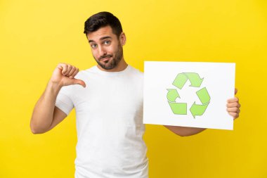 Sarı arka planda izole edilmiş yakışıklı beyaz bir adam elinde gurur verici bir jestle geri dönüşüm simgesiyle bir pankart tutuyor.