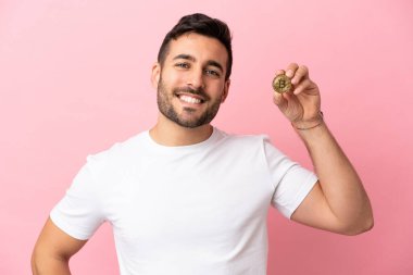 Elinde pembe arka planda izole edilmiş bir Bitcoin tutan genç adam kolları kalçasında poz veriyor ve gülümsüyor