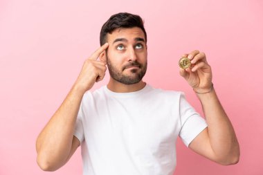 Elinde pembe arka planda izole edilmiş bir Bitcoin tutan genç adamın şüpheleri ve düşünceleri var.