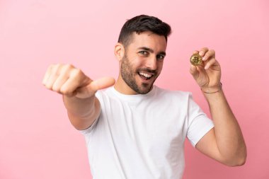 Elinde pembe arka planda izole edilmiş Bitcoin tutan genç adam iyi niyet gösterisi yapıyor.