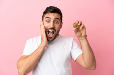 Elinde pembe arka planda, şaşırmış ve şok olmuş bir yüz ifadesi olan Bitcoin tutan genç adam.