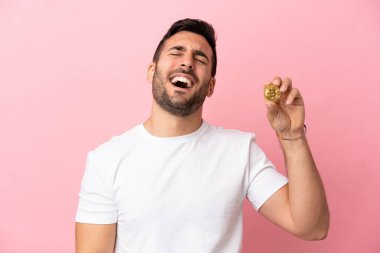 Pembe arka planda izole edilmiş Bitcoin tutan genç adam gülüyor.