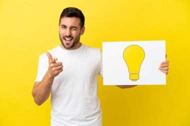 Sarı arka planda izole edilmiş yakışıklı beyaz adam elinde ampul simgesi olan bir levha tutuyor ve ön tarafı işaret ediyor.