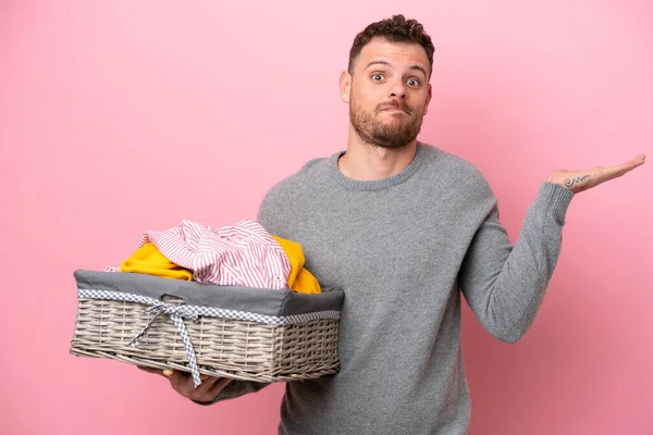 Genç Brezilyalı bir adam pembe arka planda izole edilmiş bir çamaşır sepetini tutarken ellerini kaldırırken şüphe duyuyor.