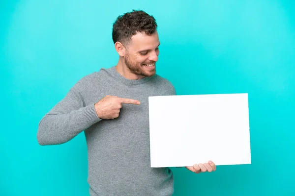拿着一张蓝色背景孤立的空标语牌的巴西年轻人 拿着一张带着快乐表情的空标语牌 指着它 — 图库照片