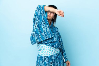 İzole edilmiş mavi arka planda kimono giyen genç bir kadın elleriyle gözlerini kapatıyor.