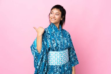 İzole edilmiş mavi arka planda kimono giyen genç bir kadın bir ürünü sunmak için kenarı işaret ediyor.