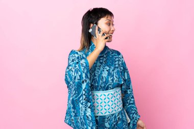 Kimono giyen genç bir kadın izole edilmiş mavi arka planda cep telefonuyla sohbet ediyor.