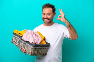 Mavi arka planda izole edilmiş çamaşır sepetini tutan orta yaşlı bir kadın parmaklarıyla tamam işareti gösteriyor.