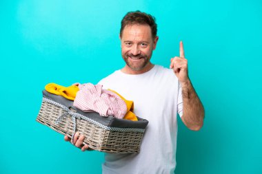 Mavi arka planda izole edilmiş çamaşır sepetini tutan orta yaşlı bir kadın en iyinin işareti olarak parmağını kaldırıyor.