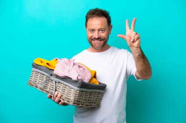 Mavi arka planda izole edilmiş çamaşır sepetini tutan orta yaşlı kadın mutlu ve parmaklarıyla üçe kadar sayıyor.