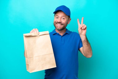 Orta yaşlı bir adam mavi arka planda izole edilmiş bir paket yiyecek alıyor gülümsüyor ve zafer işareti gösteriyor.