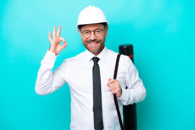 Middle age architect man with helmet and holding blueprints isolated on blue background showing ok sign with fingers