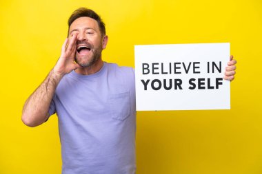 Middle age caucasian man isolated on yellow background holding a placard with text Believe In Your Self and shouting