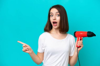 Mavi arka planda izole edilmiş saç kurutma makinesini tutan genç Ukraynalı şaşırmış ve parmağıyla kenarı işaret ediyor.