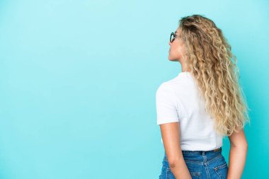 Kıvırcık saçlı kız mavi arka planda izole edilmiş ve arkasına bakıyor.