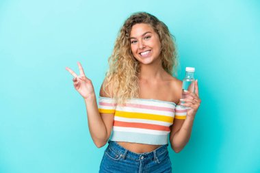 Mavi arka planda bir şişe suyu olan genç sarışın kadın gülümsüyor ve zafer işareti gösteriyor.