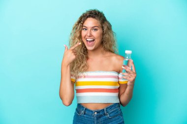 Mavi arka planda izole edilmiş su şişesi olan genç sarışın bir kadın baş parmak işareti yapıyor.