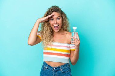 Mavi arka planda bir şişe suyu olan sarışın genç bir kadın. Sürpriz bir ifadeyle.