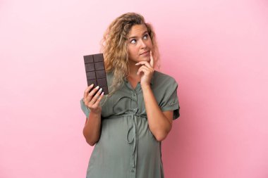 Kıvırcık saçlı kız pembe arka planda izole edilmiş hamile ve elinde çikolata tutarken şüpheleri var.