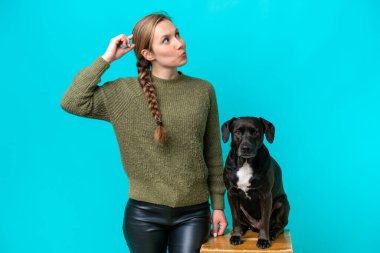 Köpeği mavi arka planda izole edilmiş genç beyaz bir kadın. Şüpheleri var ve kafası karışmış bir yüz ifadesi var.