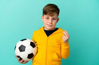 Mavi arka planda izole bir şekilde futbol oynayan kızıl saçlı çocuk para hareketi yapıyor.