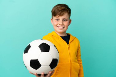 Küçük kızıl saçlı çocuk mavi arka planda futbol topuyla izole edilmiş.