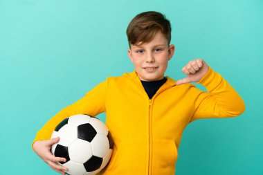 Küçük kızıl saçlı çocuk mavi arka planda izole edilmiş futbol topuyla ve kendisiyle gurur duyuyor.
