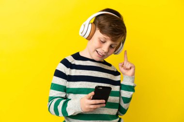 Küçük kızıl saçlı çocuk sarı arka planda izole edilmiş. Cep telefonuyla müzik dinliyor ve şarkı söylüyor.