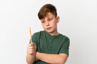 Küçük kızıl saçlı çocuk, elinde diş fırçası olan üzgün bir ifadeyle beyaz arka planda izole edilmiş.