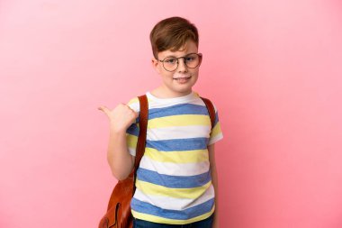 Pembe arka planda izole edilmiş küçük kızıl saçlı çocuk bir ürünü sunmak için kenarı işaret ediyor.