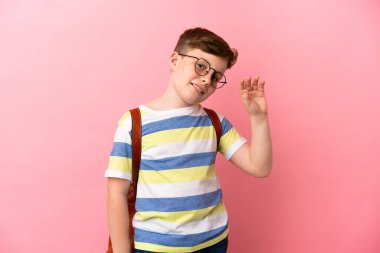 Küçük kızıl saçlı beyaz çocuk pembe arka planda izole edilmiş mutlu bir ifadeyle selam veriyor.