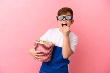 Küçük kızıl saçlı çocuk pembe arka planda 3 boyutlu gözlüklü ve elinde patlamış mısır dolu bir kova tutuyor.