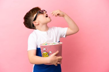 Küçük kızıl saçlı çocuk pembe arka planda 3 boyutlu gözlüklü ve elinde patlamış mısır dolu bir kova tutuyor.