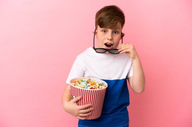 Pembe arka planda izole edilmiş küçük kızıl saçlı çocuk 3D gözlüklü ve elinde koca bir kova patlamış mısırla şaşırdı.