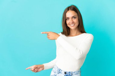 İzole edilmiş mavi arka planda genç bir kadın parmak göstererek bir ürün sunuyor.
