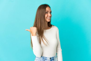İzole edilmiş mavi arka planda genç bir kadın bir ürünü sunmak için kenarı işaret ediyor.