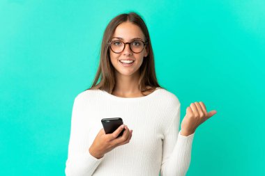 Young woman over isolated blue background using mobile phone and pointing to the lateral